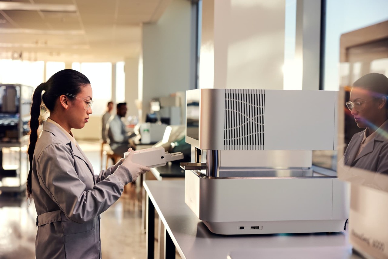Female scientist, side view, standing in front of NextSeq 1000/2000, inserting cartridge, two other scientists blurry in the background