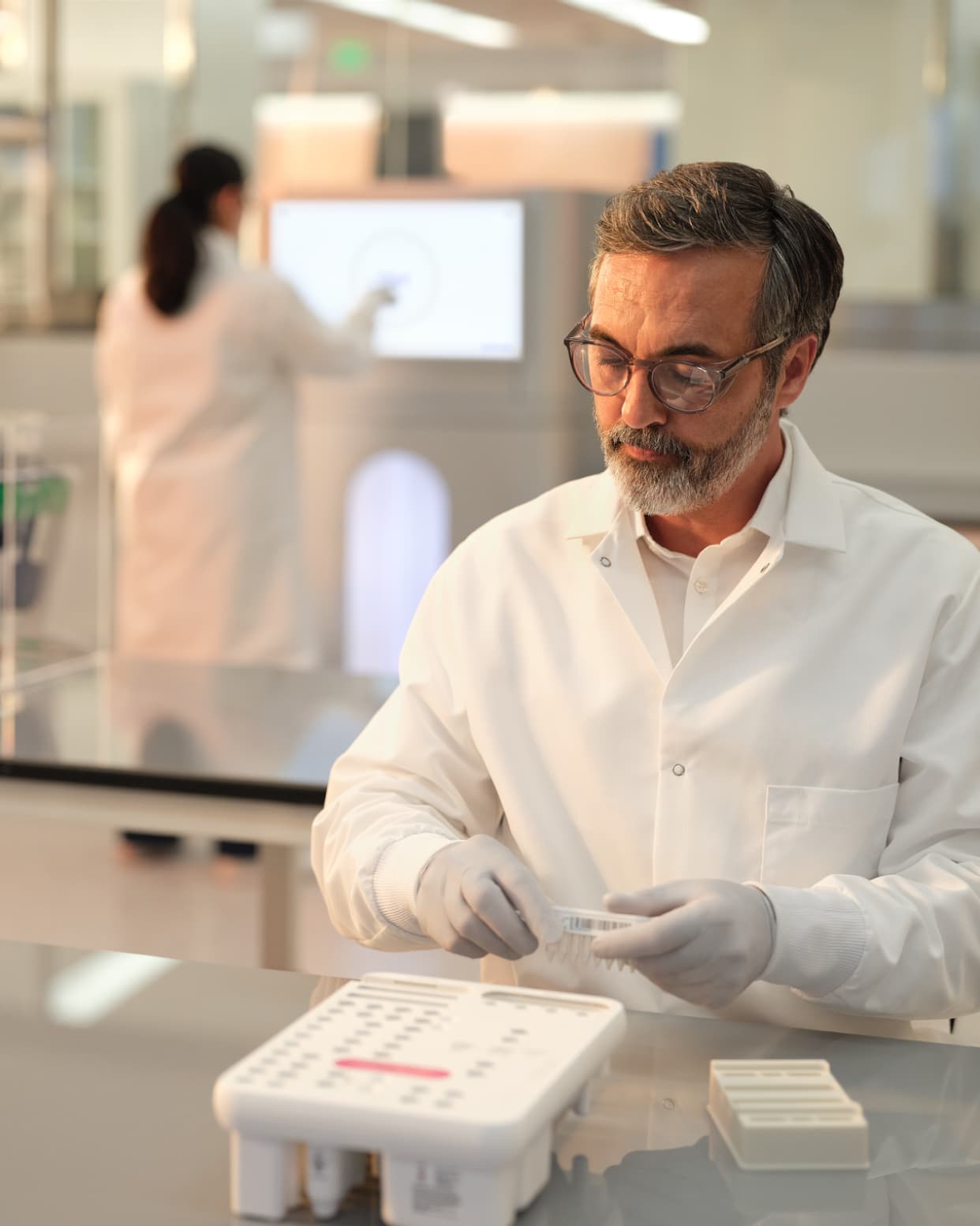 Front view of a scientist loading an 8 lane library tube strip into a reagent cartridge for a NovaSeq X run; blurry image of two female scientists interacting with a touch screen on a NovaSeq X in the background.  