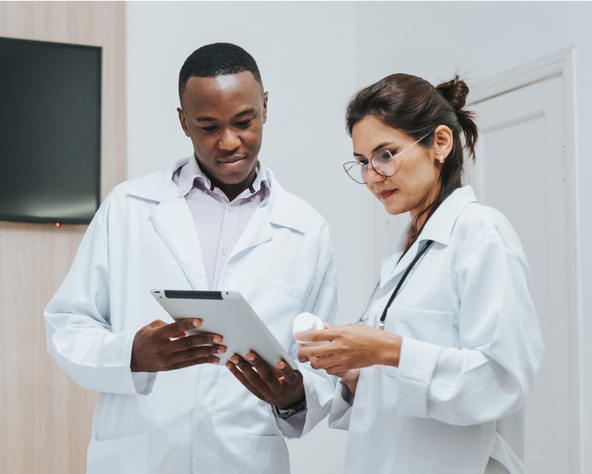 Male and female healthcare providers talking in office, looking down at a the tablet, man holding tablet and woman holding pill bottle. Patients blurry in the foreground.