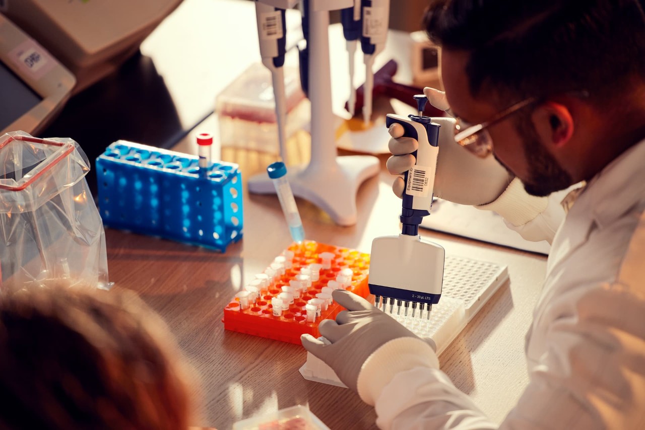 Male scientist, back side view, pipetting into 8 lane plate in wet lab, plates and tubes in background, other scientist blurry in the foreground