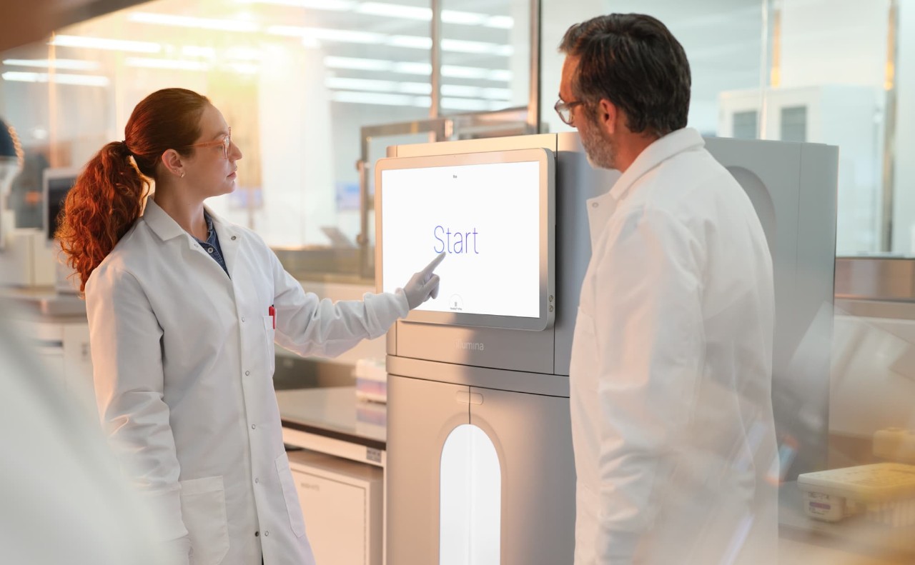 Male and female scientists touching start screen to begin run on the NovaSeq X. Consumables on lab bench next to the instrument and close up blurry image of a scientist walking in the foreground.