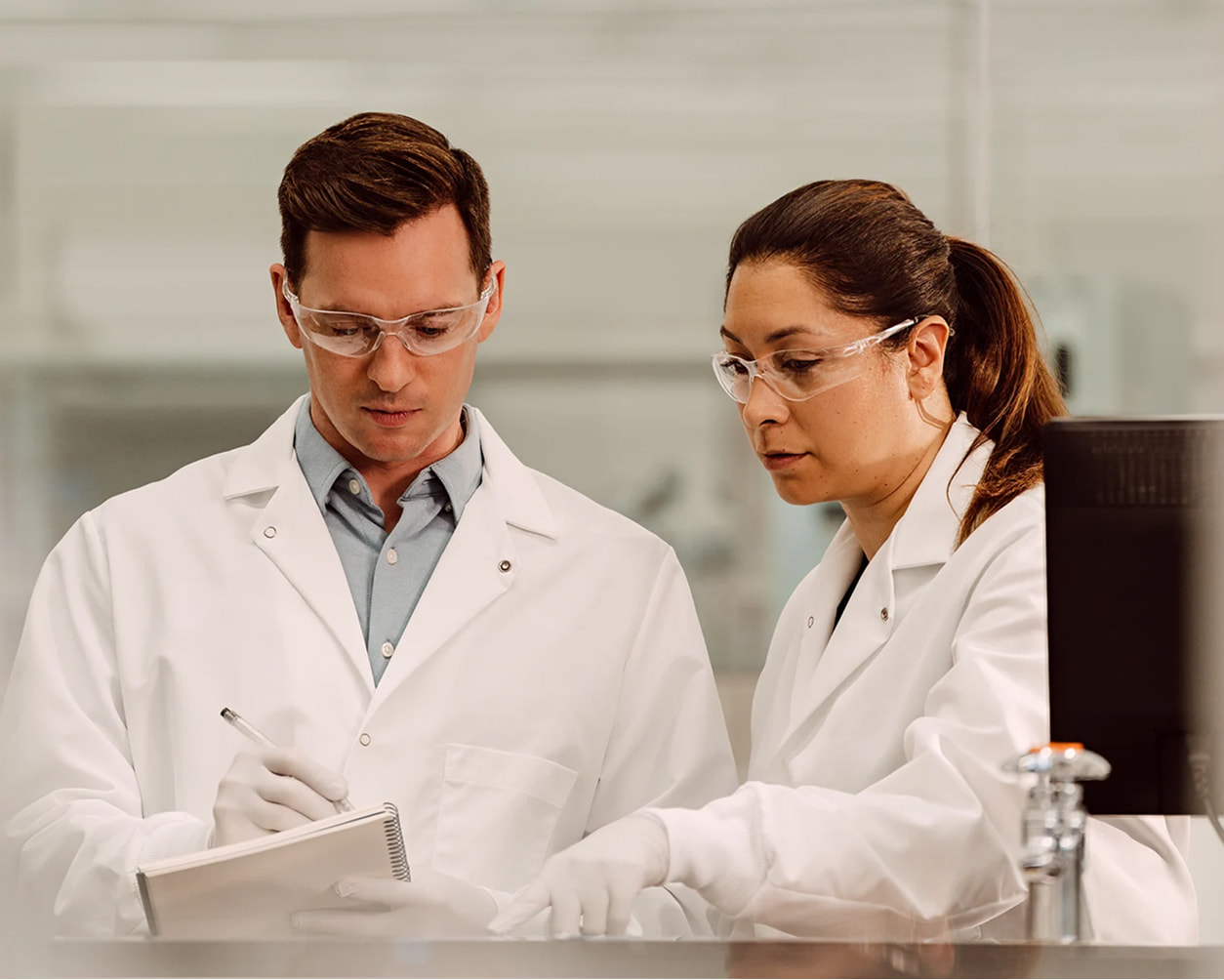 Two scientists, male and female, using a pen to write on a notepad while analyzing on a monitor, not visible, in a lab setting.
