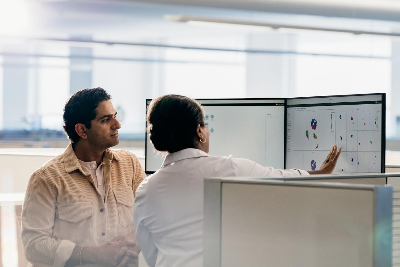 Profile view of a man and back view of a woman in an office, analyzing and discussing data on dual screens.